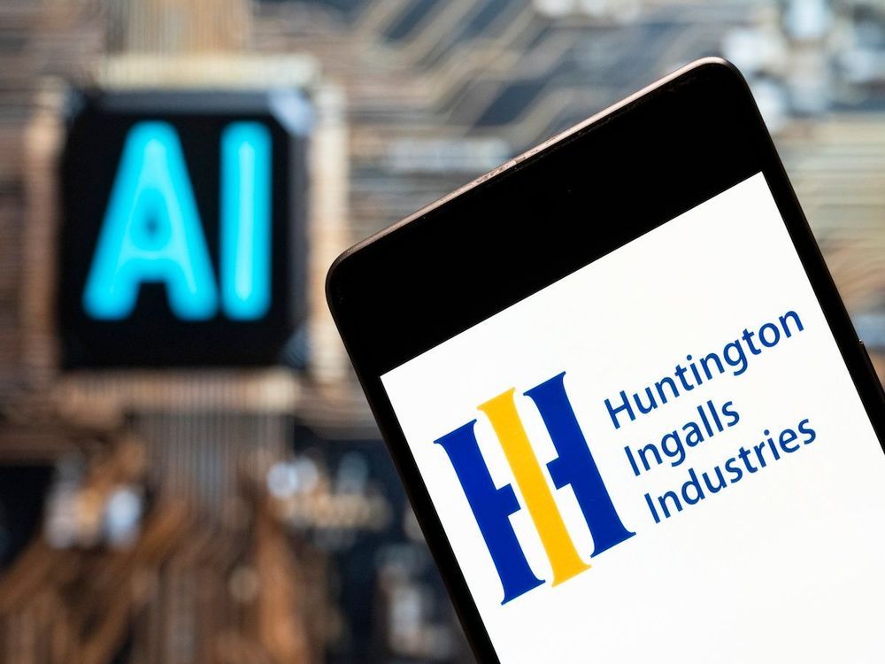In this photo illustration, America's largest military shipbuilding company, Huntington Ingalls Industries' logo seen displayed on a smartphone with an Artificial intelligence chip and symbol in the background. | Image Source: Getty Images
